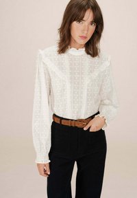 Blusa bianca e texturizzata con colletto e maniche arricciate, caratterizzata da motivi floreali. Abbinata a pantaloni scuri e una cintura marrone.