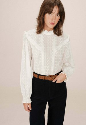 Blusa bianca e texturizzata con colletto e maniche arricciate, caratterizzata da motivi floreali. Abbinata a pantaloni scuri e una cintura marrone.