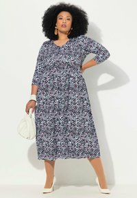 Ein knielanges, gemustertes Kleid mit roten, weißen und blauen Formen, mit rundem Ausschnitt und dreiviertel Ärmeln. Trägt weiße Absatzschuhe.