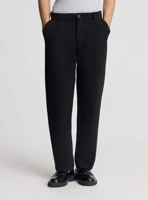 Persona che indossa pantaloni neri plissettati, scarpe di pelle nere e una camicia grigio chiaro, in piedi con le mani in tasca.