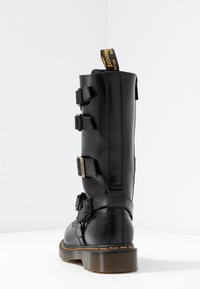 Botte haute noire en cuir brillant, avec trois sangles ajustables, coutures renforcées et semelle en caoutchouc texturée avec un accent jaune.