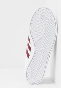 Vit atletiksko med vinröd tre-stripesdesign, gummisula med zigzag-mönster och diskret logotyp på sidan.