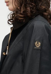 Γυναίκα με σγουρά μαλλιά που φοράει μαύρο μπουφάν Belstaff με χρυσό φερμουάρ και λογότυπο στο πάνω μέρος του μανικιού.