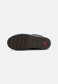 Semelle de botte en caoutchouc noir avec surface texturée, présentant des rainures profondes pour une adhérence optimale et un logo circulaire rouge près du talon.