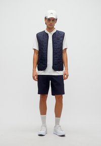 Νέος άνδρας με λευκό καπέλο, λευκό πόλο, σκούρο μπλε καπιτονέ γιλέκο, σκούρο μπλε σορτς, λευκές κάλτσες και λευκά αθλητικά παπούτσια, στεκόμενος μπροστά σε μονόχρωμο φόντο.