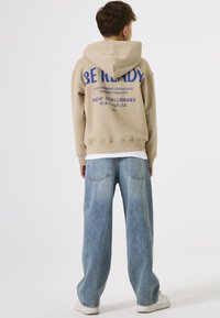Beige Kapuzenpullover mit einem großen blauen Textdruck auf dem Rücken, kombiniert mit hellblauen weiten Jeans und weißen Sneakers. Glatter Stoff, lockerer Schnitt.