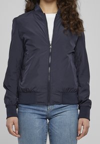Femme portant une veste bomber bleu marine zippée sur une chemise blanche et un jean bleu clair, debout devant un fond uni.