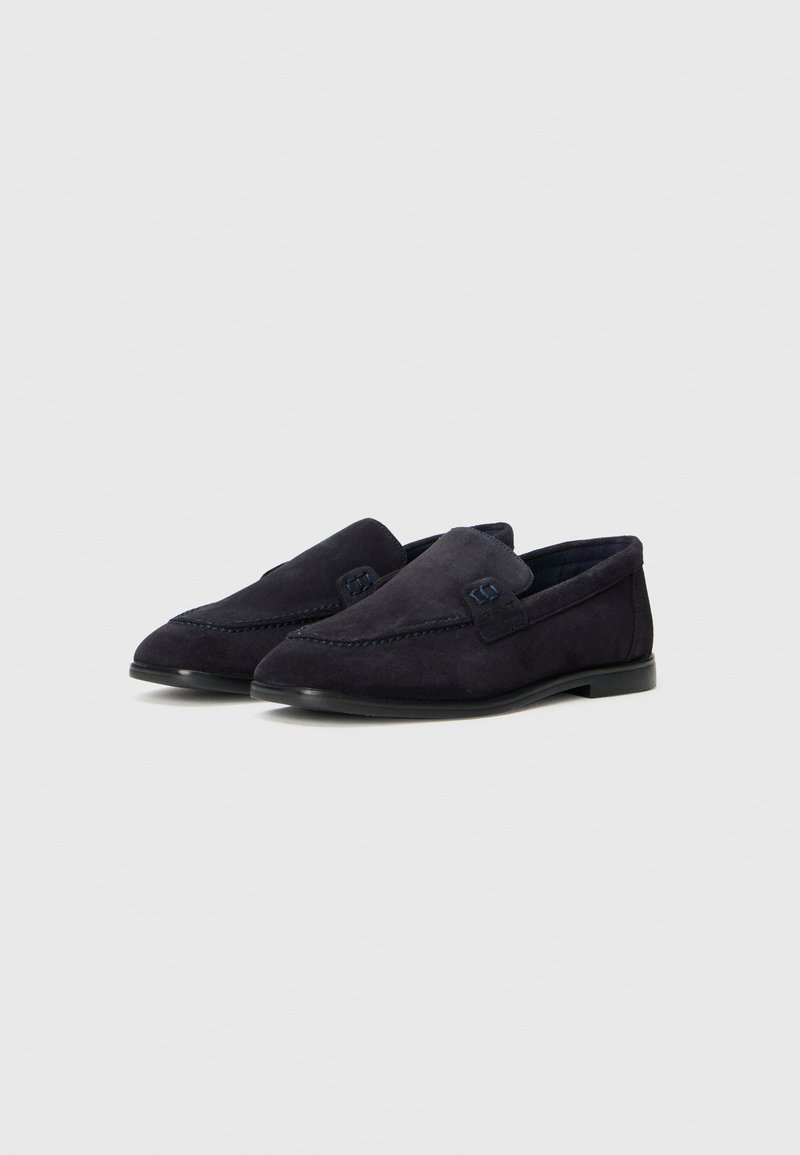 Un paio di mocassini slip-on in suede blu scuro con dettagli cuciti e suole in gomma nera basse su uno sfondo bianco semplice.