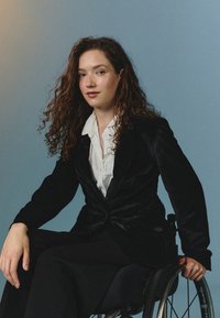 Blazer de terciopelo negro sobre una camisa blanca, sentado en una silla de ruedas. Cabello rizado, fondo neutro, centrado en las texturas y colores de la prenda.