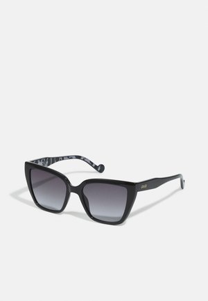 Black oversized sunglasses with a gradient lens. Plastic frame with a glossy finish. Inner temple features a patterned design. Brand name printed on the side.