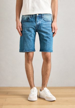 Τζιν σορτς σε ανοιχτό μπλε χρώμα, ευθεία γραμμή, με φθαρμένο στρίφωμα και σχεδίαση πέντε τσέπες. Φοριέται με λευκά αθλητικά παπούτσια πάνω σε ξύλινο πάτωμα.