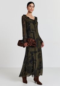 Robe longue en voile vert olive avec des motifs noirs complexes, décolleté à volants et manches longues ; associée à une pochette marron et des bottes au genou.