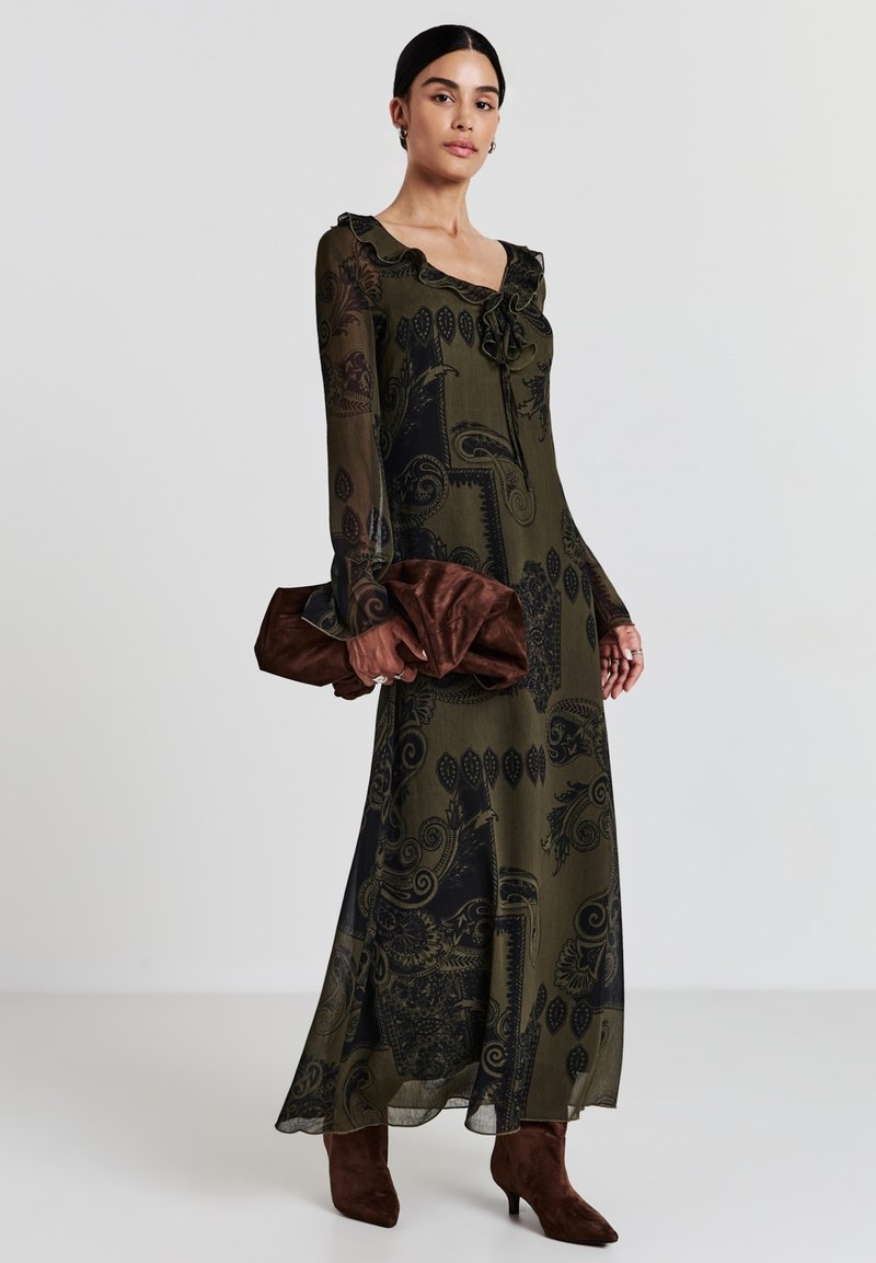 Robe longue en voile vert olive avec des motifs noirs complexes, décolleté à volants et manches longues ; associée à une pochette marron et des bottes au genou.