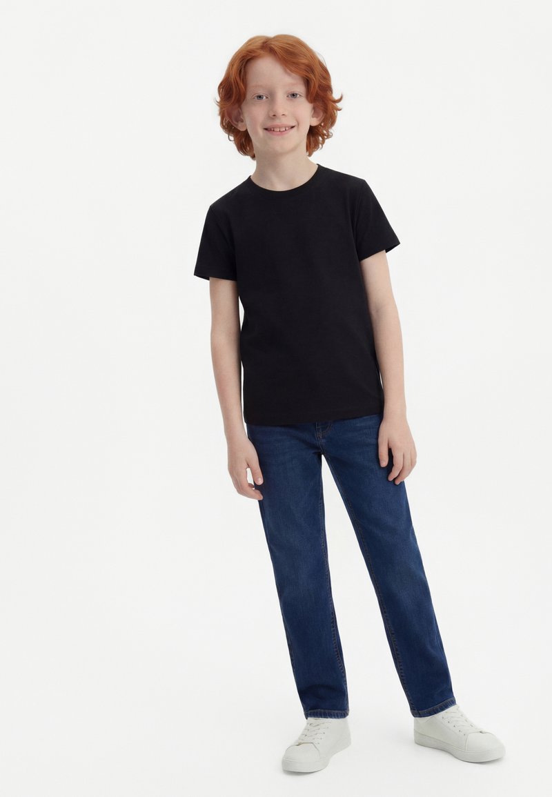 Rothaariges Kind steht, trägt ein schwarzes kurzärmeliges T-Shirt, blaue Jeans und weiße Turnschuhe, lächelt vor einem schlichten weißen Hintergrund.