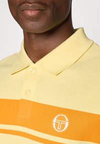 Geel poloshirt met oranje strepen, voorzien van een klassieke kraag en drie knopen. Een wit logo is zichtbaar op de linkerkant van de borst.