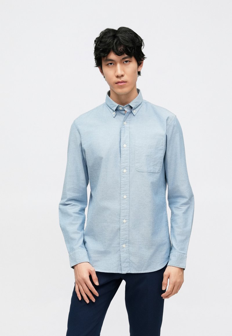 Camisa azul clara de manga comprida com colarinho com botões, bolso no peito e fecho de botões, feita de um tecido texturizado.