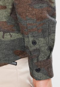 Chemise à motif camouflage avec des couleurs vertes, marron et grises. Dotée d'un poignet boutonné et d'un tissu texturé. Étiquette de la marque visible sur le côté.