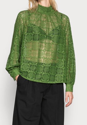 Femme portant un chemisier en dentelle verte transparente à col montant avec manches longues et pantalon noir, main dans la poche, sur fond uni.