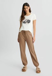 Crème t-shirt met zwart bloemenontwerp en rode strepen; gecombineerd met bruine joggerbroeken met een elastische tailleband en enkelsluiting.