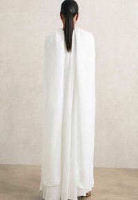 Cape fluide blanche en tissu léger, tombant élégamment des épaules jusqu'au sol, avec une texture lisse et douce et des détails minimaux.