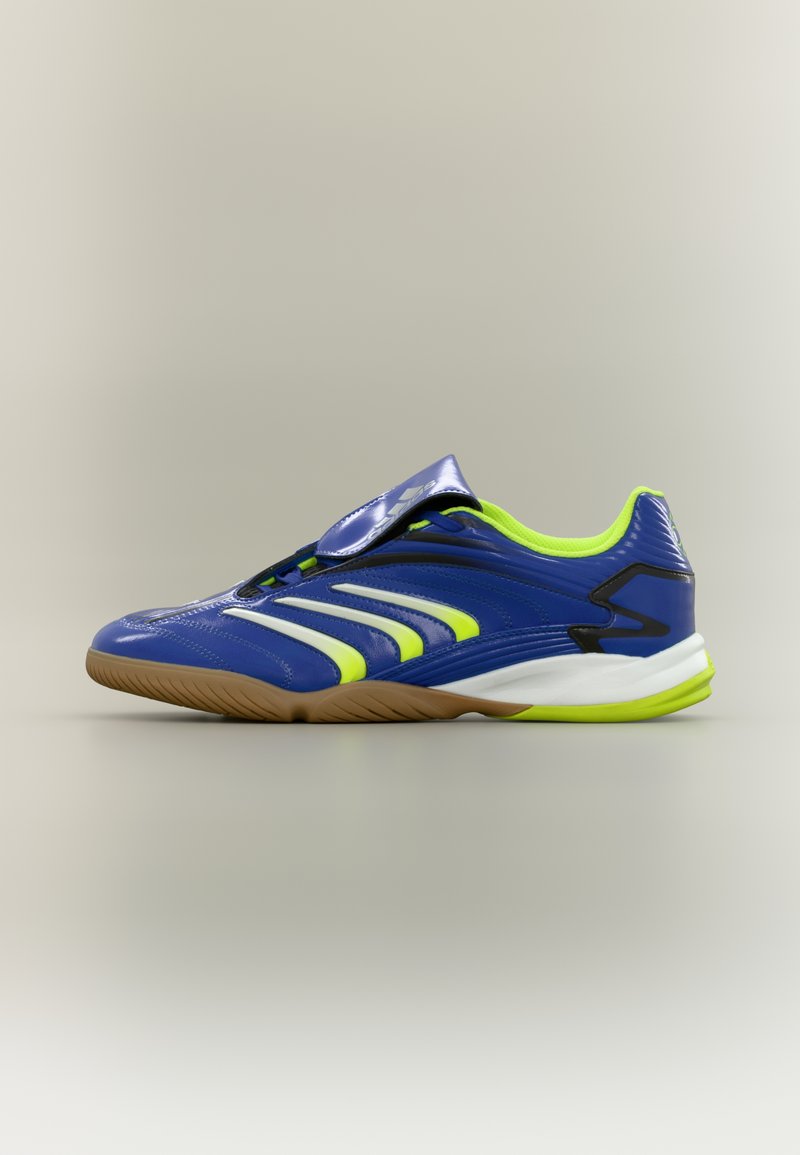 Chaussure de sport bleue avec des accents vert néon, une semelle marron et une bride à velcro sur fond beige, vue de profil.