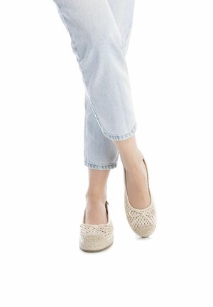 Zapatos slip-on trenzados color beige con punta redondeada, que presentan un lazo decorativo, combinados con jeans recortados de color azul claro. Textura suave y diseño transpirable.