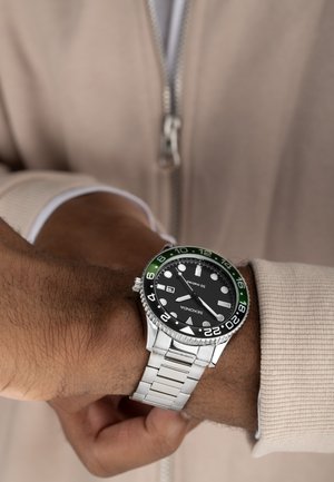 Montre en métal argenté avec un cadran noir et une lunette verte, dotée de marqueurs luminescents, d'un guichet de date et d'un bracelet en acier inoxydable.