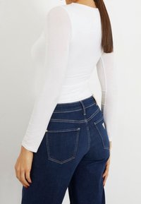 Woman wearing a fitted white long-sleeve top and high-waisted dark blue denim jeans, shown from the back and waist up.