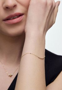 Złota bransoletka o delikatnym wzorze łańcuszka, z małymi okrągłymi akcentami w kolorze czarnym i złotym. Noszona na nadgarstku o jasnej karnacji.