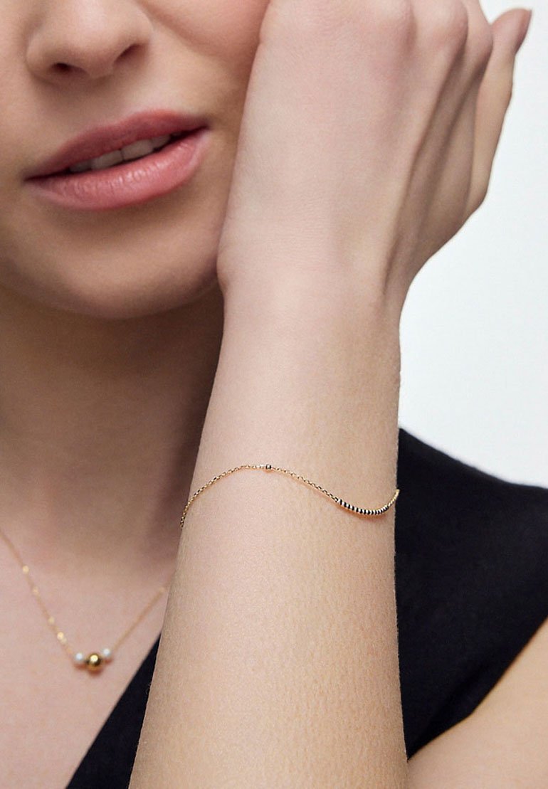 Złota bransoletka o delikatnym wzorze łańcuszka, z małymi okrągłymi akcentami w kolorze czarnym i złotym. Noszona na nadgarstku o jasnej karnacji.