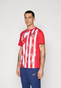 Piros sportpóló, fehér függőleges csíkokkal és texturált mintával, a mellkason egy logóval. Kék nadrággal viselve.