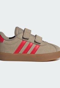 Smeđe antilop tenisice s dva crvena pruga i čičak trakama. Potplat je od smeđeg gume s teksturiranom površinom za bolju prijanjanje.