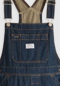 Mørkeblå denim overaller med justerbare stropper, sølvfarget metall, oransje sømmer og en baklomme med et merke.
