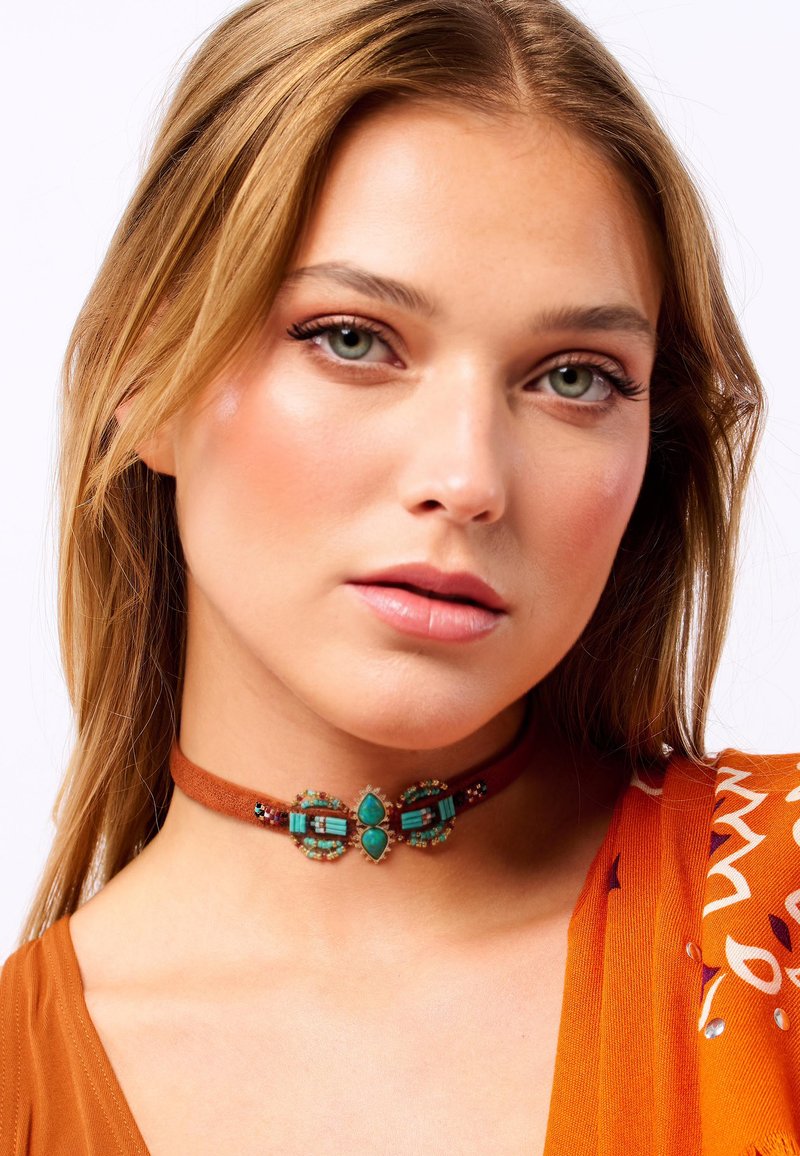 Choker en daim marron avec des accents en turquoise et en métal, présentant une pièce centrale décorative et des détails perlés, porté avec un haut à motifs.