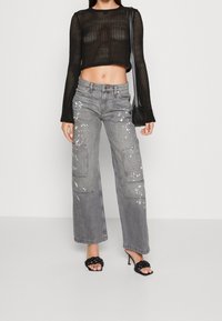 Haut en tricot noir court avec manches longues, associé à un pantalon cargo gris à jambes larges comportant des éclaboussures de peinture et de grandes poches latérales. Sandales à talons noires.