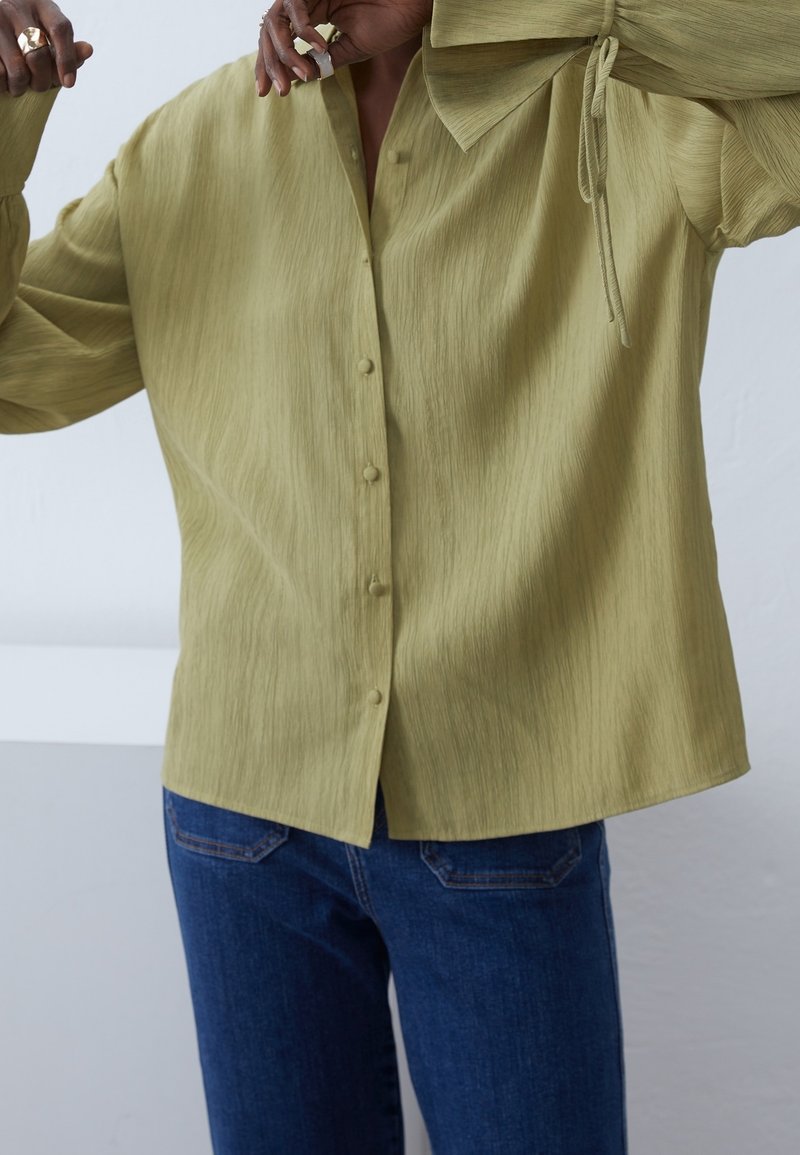 Personne portant une chemise texturée vert clair avec boutons et grand col, et un jean bleu foncé, levant les bras près du visage.