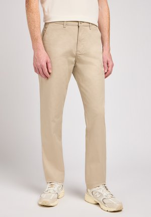 Chinos beige con corte recto, confeccionados en tela de algodón. Incluyen bolsillos clásicos y se llevan con zapatillas blancas y beige.