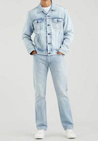 Chaqueta de mezclilla azul claro con cierre de botones y dos bolsillos en el pecho, combinada con jeans azul claro. Zapatillas blancas completan el look.