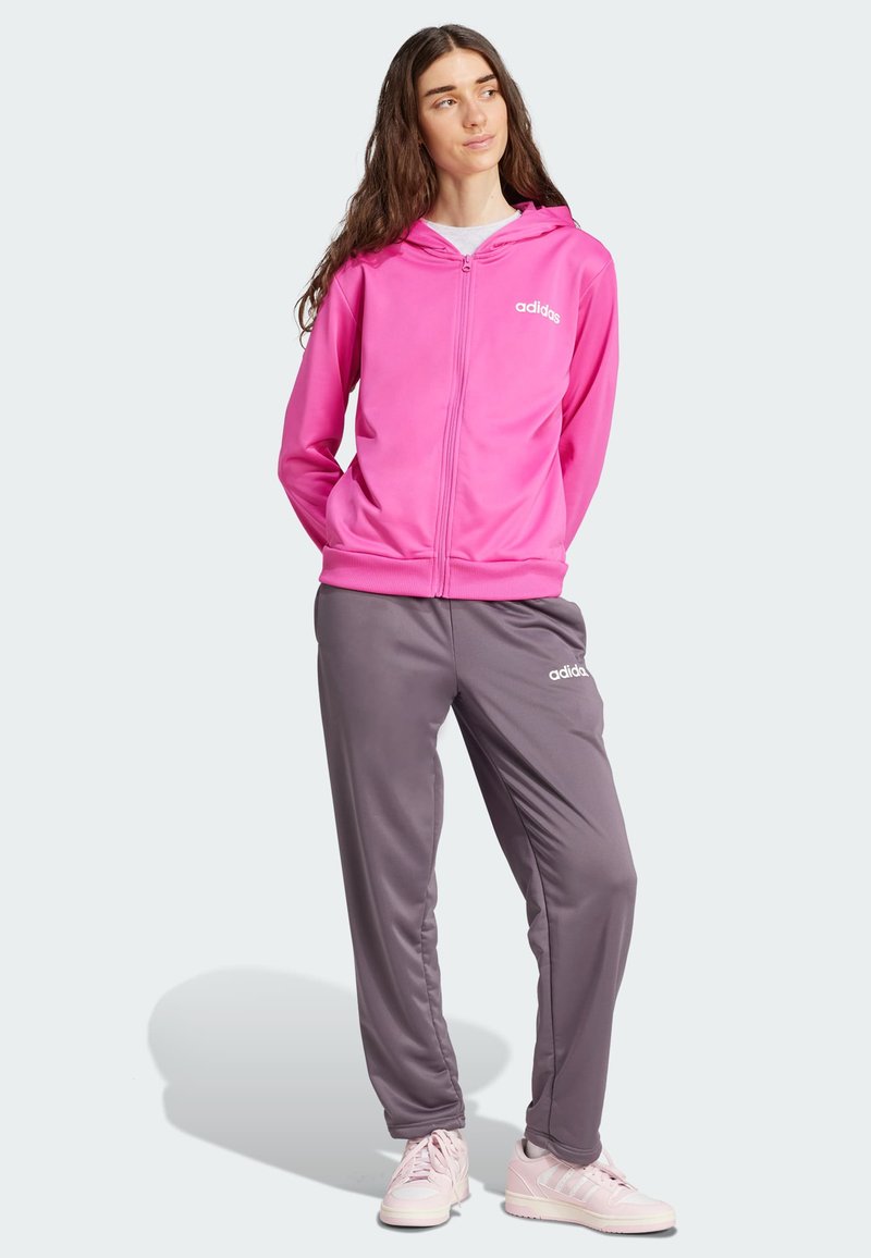 Sudadera rosa con cremallera y capucha, combinada con pantalones de chándal grises que tienen un logo de Adidas. El conjunto incluye zapatillas deportivas rosas.