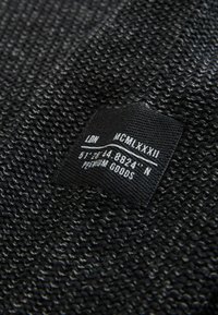 Czarna tkana tkanina o teksturowanym wykończeniu. Czarna metka przedstawia tekst, w tym "LON", "MCMLXXXII" oraz współrzędne, z napisem "PREMIUM GOODS".