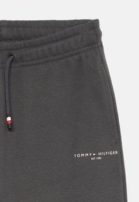 Šedé tepláky s elastickým pásom na šnúrku, s bielym vyšitým logom a malým červeným a modrým akcentom na konci šnúrky.