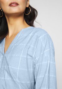 Haut en lin bleu clair avec un col en V et de fines rayures blanches verticales. Caractérisé par des manches amples et un tissu doux et texturé.