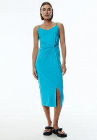 Ein türkisfarbenes Midikleid mit einem geknoteten Detail an der Taille, dünnen Trägern und einem seitlichen Schlitz, aus einem glatten, dehnbaren Stoff.