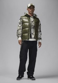 Vadderad olivgrön väst över en grafisk sweatshirt, i kombination med mörka cargobyxor och svarta sneakers. Tillbehör inkluderar en keps med en logotyp.