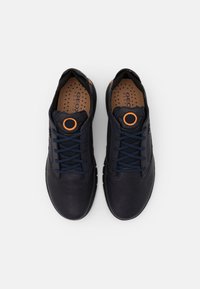 Une paire de baskets à lacets bleu marine foncé avec un détail circulaire orange sur la languette et des semelles perforées beiges, vue de dessus.