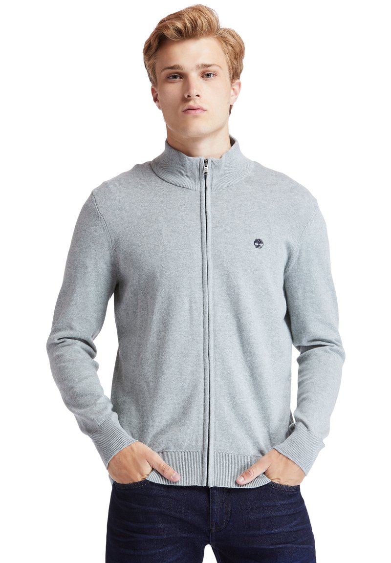 Sivá zip-svetra s visokim ovratnikom, rebrastimi manžetami in spodnjim delom. Ima manjši logotip na prsih. Tkanina se zdi mehka in teksturirana.
