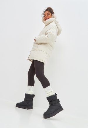 Manteau en duvet crème à capuche, leggings noirs et bottes isolées noires avec une boucle. Des chaussettes blanches en tricot ajoutent de la texture.