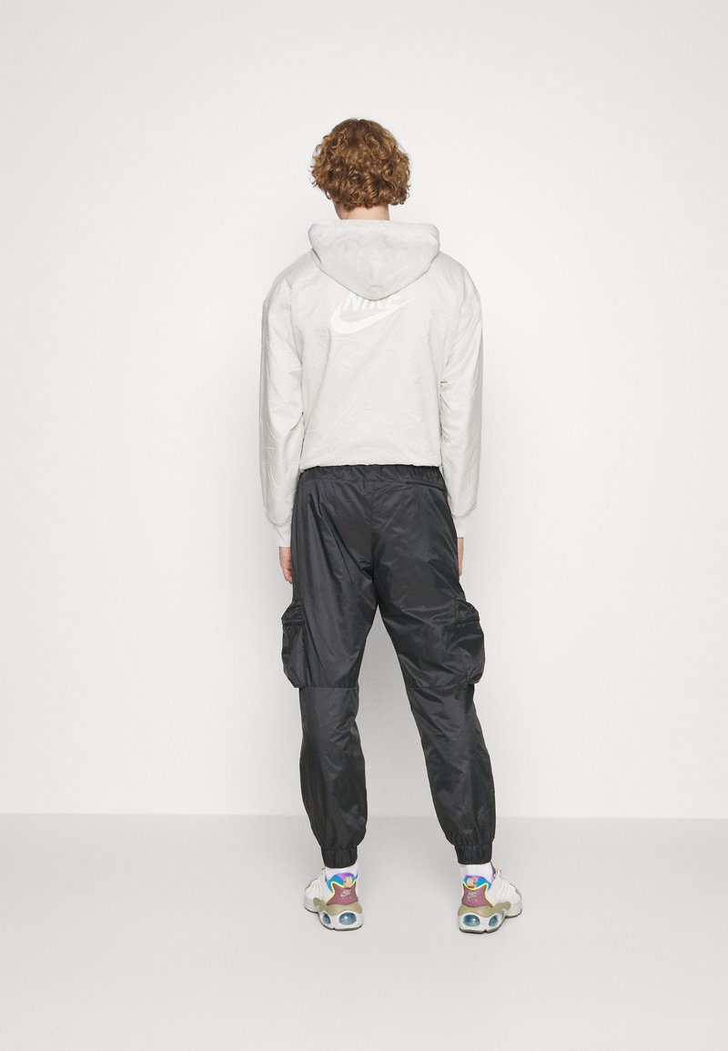 Person mit lockigem Haar, die eine weiße Nike-Hoodie-Jacke, schwarze Cargohosen und weiße Sneakers trägt und vor einer schlichten weißen Wand steht.