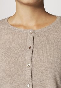 Beige ribbstickad kofta med rund halsringning. Tre knappar i varierande nyanser av brunt och gräddvitt. Mjuk textur och figursydd design.