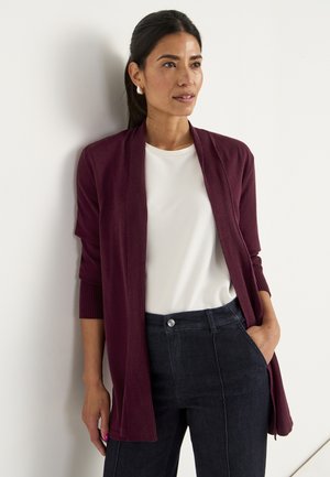 Vrouw met donker haar draagt een bordeauxrode cardigan, witte top en donkere high-waisted broek, staand tegen een witte muur, met een hand in haar pocket.